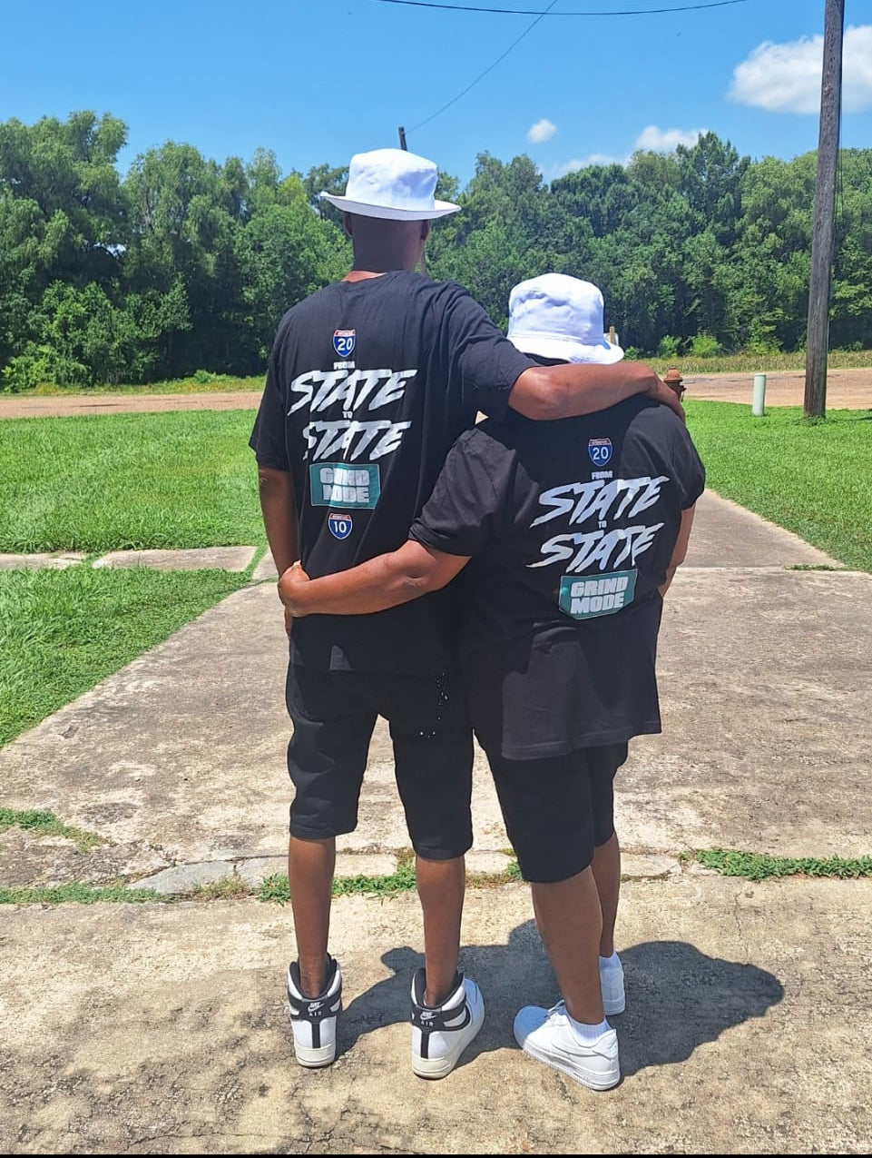 Two people wearing matching black t-shirts with 'STATE' text, standing outdoors on a sunny day.